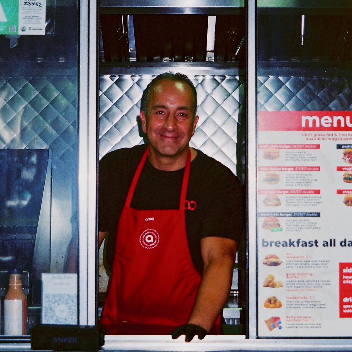 A smiling man wearing a red apron over a black t-shirt stands behind a food service counter with a menu board on the right, in Los Angeles