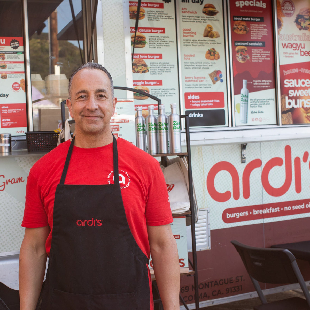 A smiling man wearing a red t-shirt and a black apron with the brand name 'ardi' stands in front of a food truck. The food truck has 'ardi' written in large red letters and displays menus for burgers, breakfast, and specials.