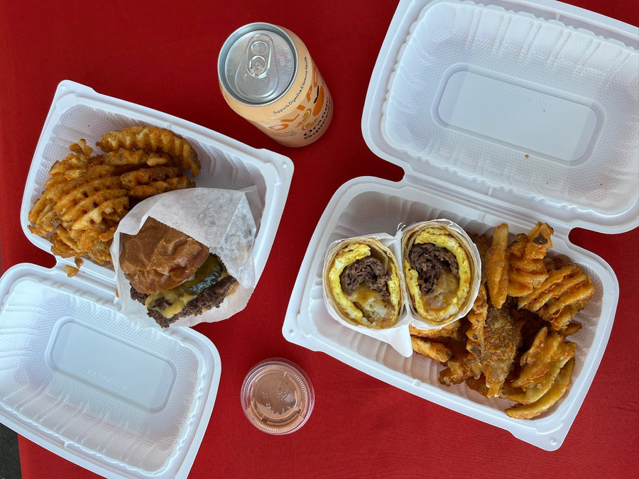 A takeout meal set on a red tablecloth, featuring two white styrofoam containers with a burger wrapped in paper, waffle fries, two egg wraps with meat, a small cup of sauce, and an orange soda can.