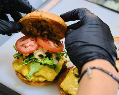 A close-up of a hand in a black glove assembling a burger with a beef patty, melted cheese, lettuce, and tomato slices on a white cutting board.